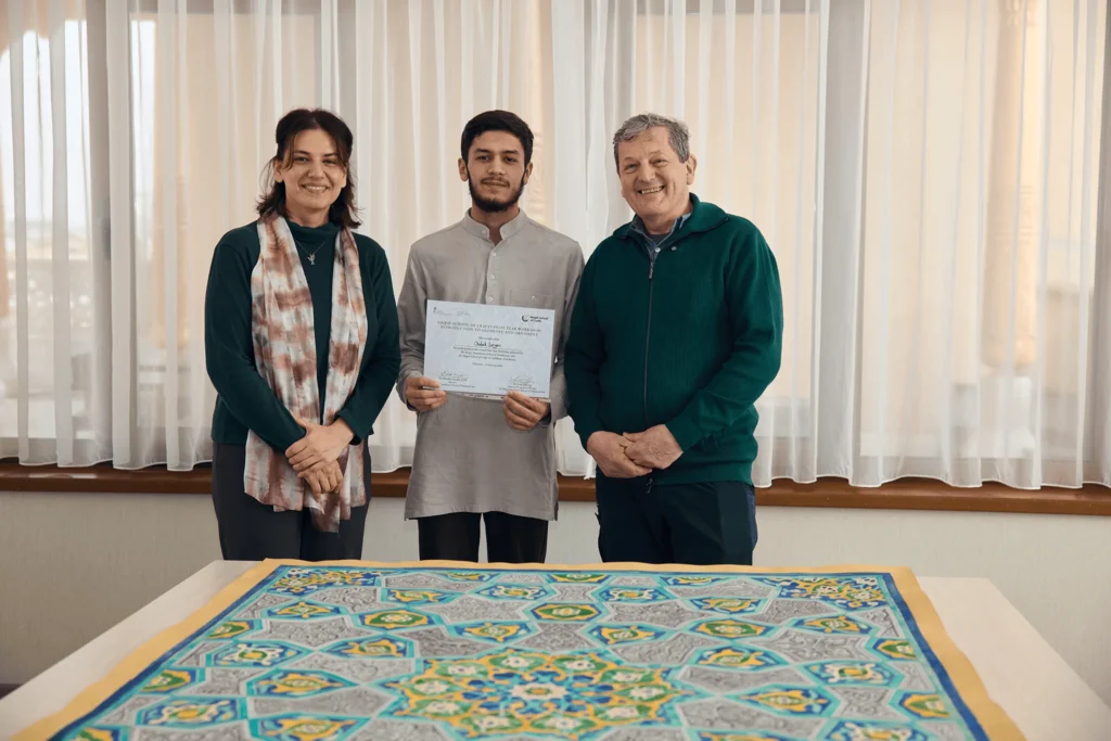 Отабек держит сертификат рядом с двумя преподавателями, на переднем плане — завершённая геометрическая работа.